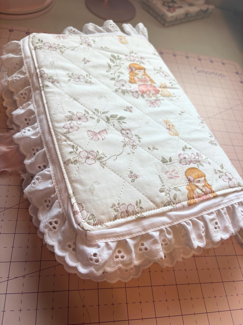 May include: A quilted, off-white journal cover with a floral and girl illustration pattern. The cover is trimmed with white eyelet lace and a satin ribbon. The journal is a soft, feminine accessory.