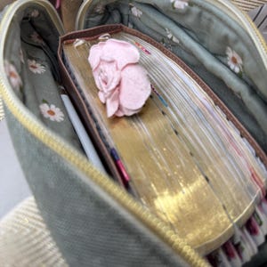 Puede incluir: Una bolsa verde con cremallera y forro floral. La bolsa est&aacute; abierta y contiene una pila de libros viejos y amarillentos con una flor rosa en la parte superior.