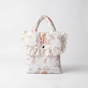 May include: A white quilted handbag with a floral pattern in pink and green. The bag features a top handle, a flap with a button closure, and a decorative lace trim. The design includes illustrations of a girl and a bunny.