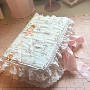 May include: A quilted journal with a floral and girl design, featuring a white eyelet lace trim and a pink satin ribbon closure. The journal's cover has a diamond pattern and a soft, feminine aesthetic.