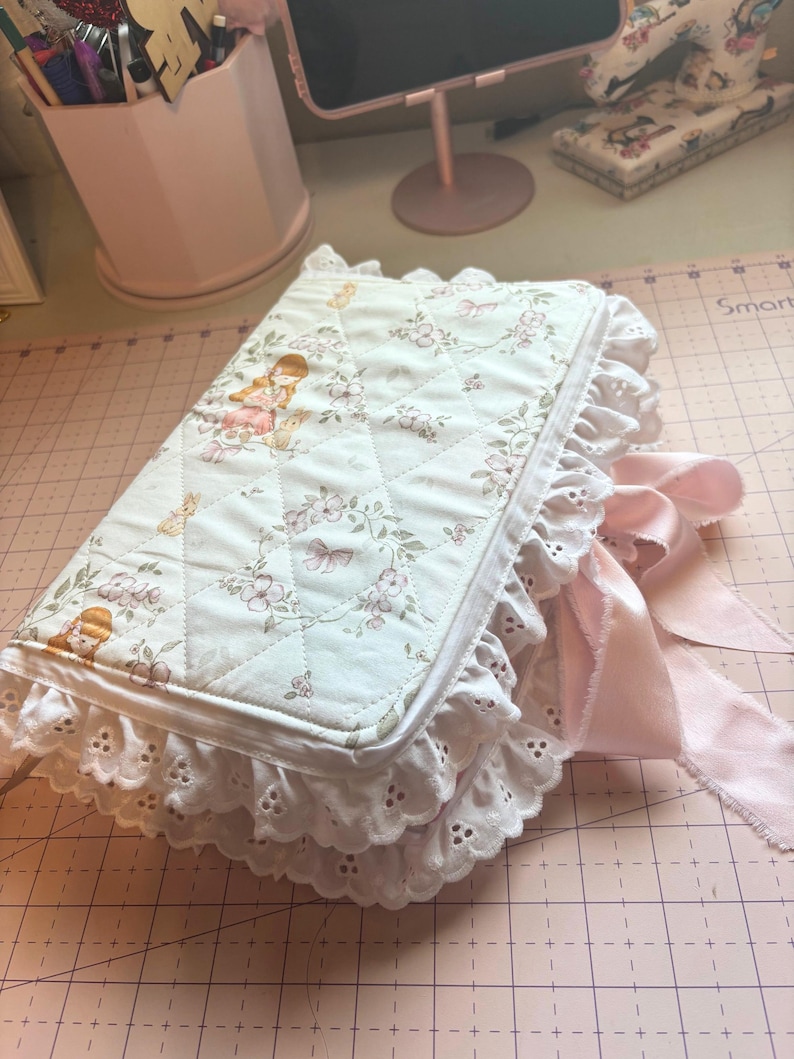 May include: A quilted journal with a floral and girl design, featuring a white eyelet lace trim and a pink satin ribbon closure. The journal's cover has a diamond pattern and a soft, feminine aesthetic.