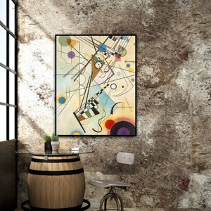 May include: An abstract art print in a black frame, featuring geometric shapes and lines in various colors, including blue, yellow, and red. The artwork hangs on a textured, neutral-toned wall. A wooden barrel table with a glass top and a metal stool are in the foreground.