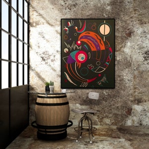 May include: Abstract art print in a black frame. The artwork features a colorful, geometric design with shapes in red, orange, purple, and green on a dark background. The image is displayed in a room with a barrel table and a metal chair.