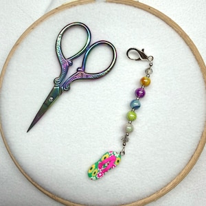 May include: Iridescent rainbow-coloured metal embroidery scissors with ornate handles alongside a silver-toned metal charm featuring a vertical row of multicoloured glass beads and a single miniature flip-flop pendant with a bright pink strap.