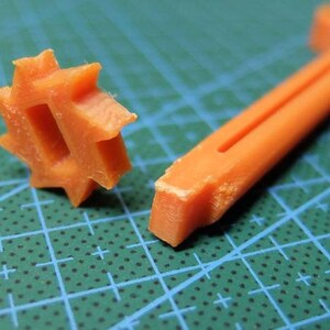 May include: Close-up of two orange plastic tools. One is a star-shaped piece with a central opening, and the other is a long, thin, L-shaped tool with a groove. The tools are on a green cutting mat.