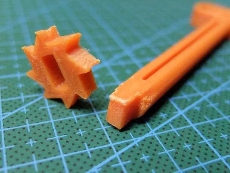 May include: Close-up of two orange plastic tools. One is a star-shaped piece with a central opening, and the other is a long, thin, L-shaped tool with a groove. The tools are on a green cutting mat.