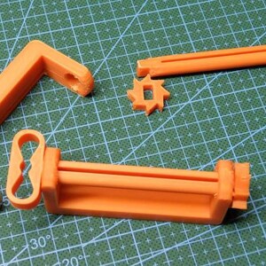 May include: Orange 3D-printed components, including an L-shaped piece, a gear-like shape, and a rectangular frame with a handle. The parts are arranged on a green cutting mat, suggesting a DIY or crafting project.