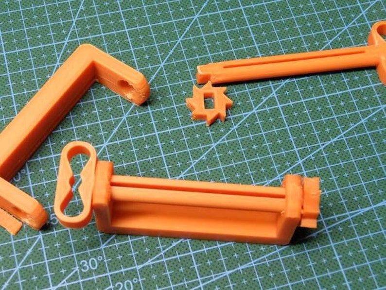 May include: Orange 3D-printed components, including an L-shaped piece, a gear-like shape, and a rectangular frame with a handle. The parts are arranged on a green cutting mat, suggesting a DIY or crafting project.