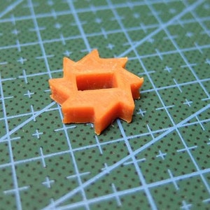 May include: An orange, star-shaped plastic component with a square hole in the center. The component has multiple points around its perimeter. It is set against a green cutting mat with blue grid lines.