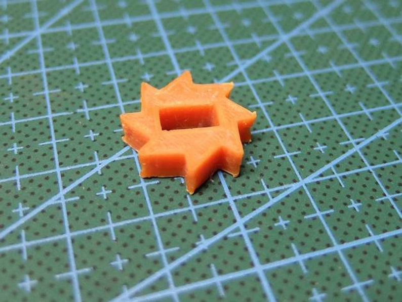 May include: An orange, star-shaped plastic component with a square hole in the center. The component has multiple points around its perimeter. It is set against a green cutting mat with blue grid lines.
