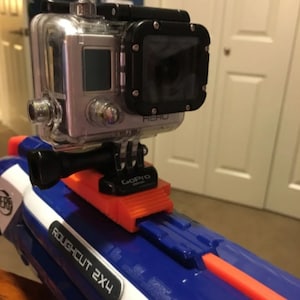 May include: A silver GoPro camera mounted on an orange and black bracket, attached to a blue and white Nerf Roughcut 2X4 blaster. The camera is in a clear protective case.