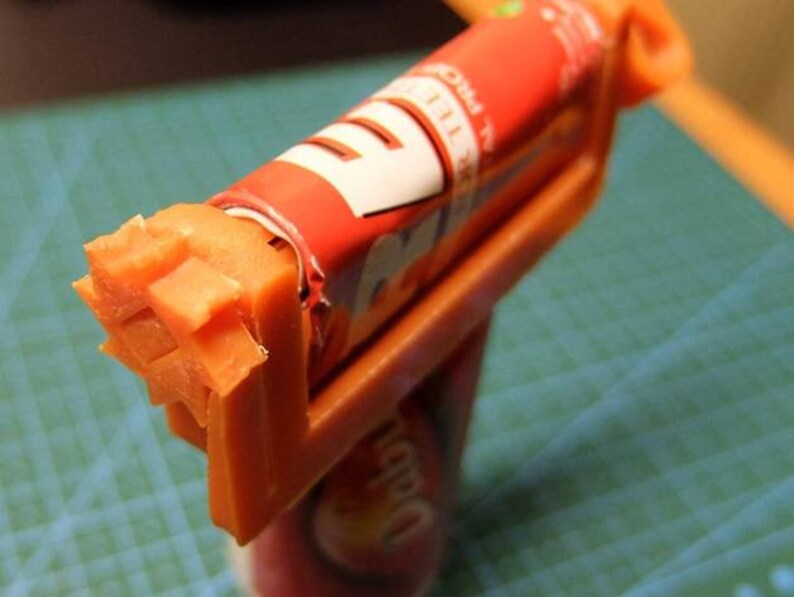 May include: An orange plastic candy cutter holding a partially unwrapped red and white candy roll. The candy has been cut into a star shape. The candy roll has the words "PRO" and "TEE" visible.