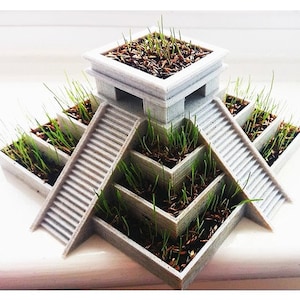 May include: A gray, tiered planter with stair-like structures, filled with soil and sprouting green grass. The design resembles a miniature architectural model or a stylized pyramid, suitable for growing small plants or herbs.