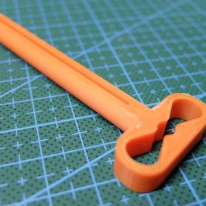 May include: An orange plastic tool with a long, rectangular handle and a looped end. The tool is designed with a central cutout in the looped section. The background is a green cutting mat.