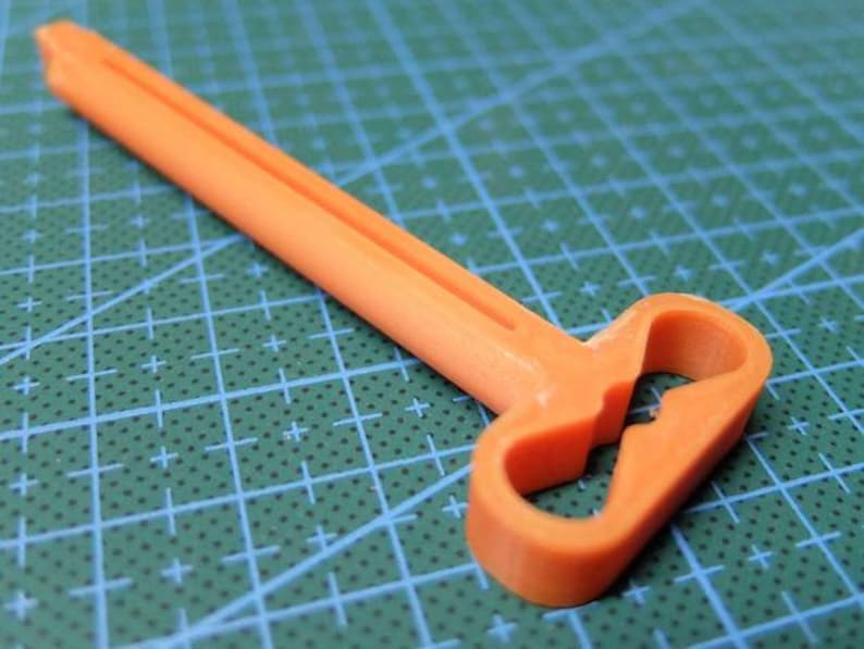 May include: An orange plastic tool with a long, rectangular handle and a looped end. The tool is designed with a central cutout in the looped section. The background is a green cutting mat.