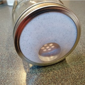 May include: Close-up of a glass spice jar with a metal lid. The lid has a white, speckled surface with a small, rounded dispenser featuring multiple holes. The jar is partially filled with a light-colored spice.