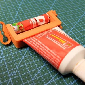 May include: An orange plastic tube squeezer with a handle, holding a red and white tube of toothpaste. Another tube of toothpaste is being squeezed. The background is a green cutting mat.