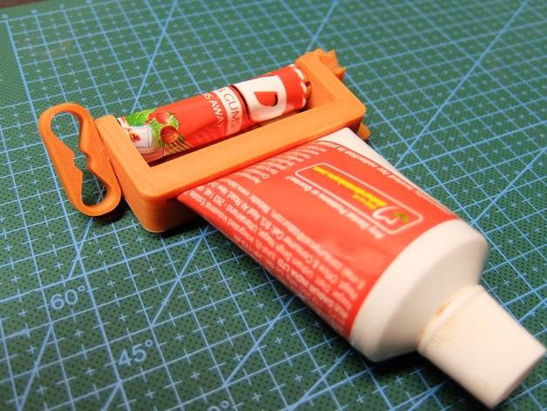 May include: An orange plastic tube squeezer with a handle, holding a red and white tube of toothpaste. Another tube of toothpaste is being squeezed. The background is a green cutting mat.
