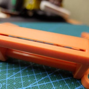 May include: Close-up of an orange plastic object with a rectangular frame and a slot. The object has a hook-like extension and is set against a green grid background. Other orange pieces are visible.