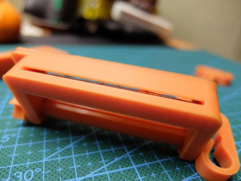 May include: Close-up of an orange plastic object with a rectangular frame and a slot. The object has a hook-like extension and is set against a green grid background. Other orange pieces are visible.