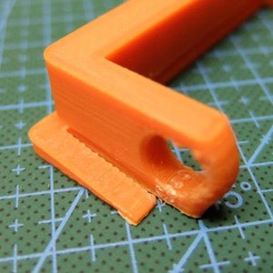 May include: Close-up of an orange plastic object with a right-angle shape. The object has a rectangular base with a slot and a curved section with a hole. The object is on a green cutting mat with white grid lines.