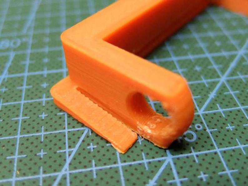 May include: Close-up of an orange plastic object with a right-angle shape. The object has a rectangular base with a slot and a curved section with a hole. The object is on a green cutting mat with white grid lines.