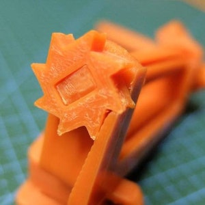 May include: Close-up of an orange plastic object with a star-shaped end and a square indentation. The object appears to be a component of a larger mechanism, possibly a tool or a machine part. The background is a teal cutting mat.