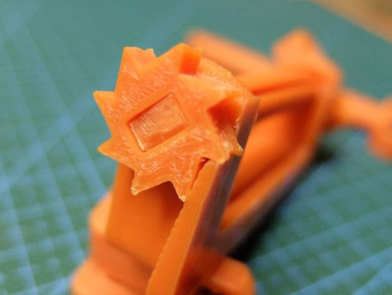 May include: Close-up of an orange plastic object with a star-shaped end and a square indentation. The object appears to be a component of a larger mechanism, possibly a tool or a machine part. The background is a teal cutting mat.