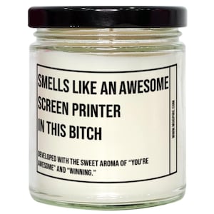 May include: A clear glass candle jar with a black lid containing white wax. The label features black text reading: SMELLS LIKE AN AWESOME SCREEN PRINTER IN THIS BITCH. Below this, it reads: DEVELOPED WITH THE SWEET AROMA OF YOU'RE AWESOME AND WINNING.