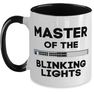 May include: White ceramic coffee mug with a black handle and black rim. The mug has the text "MASTER OF THE BLINKING LIGHTS" in black text. A graphic of a network switch with blinking lights is in the middle of the mug.