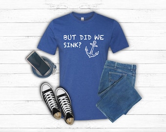 May include: Blue t-shirt with the text "BUT DID WE SINK?" and an anchor graphic. The shirt is paired with blue jeans and black and white sneakers. A smartphone and a blue cord are also in the image.