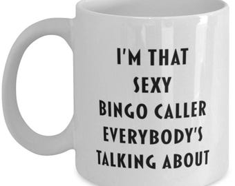 May include: A white ceramic mug with a C-shaped handle featuring the black printed text I'M THAT SEXY BINGO CALLER EVERYBODY'S TALKING ABOUT centred on the front. The mug has a smooth, glossy finish and a standard cylindrical shape for hot beverages.