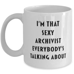 Könnte beinhalten: Ein weißer Keramikbecher mit einem Henkel auf der linken Seite. Auf der Vorderseite des Bechers befindet sich ein schwarzer Schriftzug: I'M THAT SEXY ARCHIVIST EVERYBODY'S TALKING ABOUT. Der Becher hat eine glatte, glänzende Oberfläche und eine zylindrische Form.