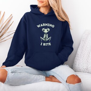 May include: Navy blue hooded sweatshirt with a front pocket. The sweatshirt features a cartoon praying mantis design with the text "WARNING I BITE" in white. The design is centered on the chest.