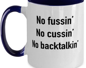 May include: A white ceramic mug with a dark blue interior and handle. The exterior features black text that reads: No fussin' No cussin' No backtalkin'. The mug has a cylindrical shape with a curved handle on the left side and a smooth finish.