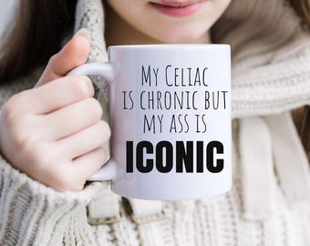 May include: A white ceramic mug featuring black printed text that reads: MY CELIAC IS CHRONIC BUT MY ASS IS ICONIC. The mug has a standard handle and a smooth, glossy finish, displaying the bold, sans-serif lettering clearly on the front surface.