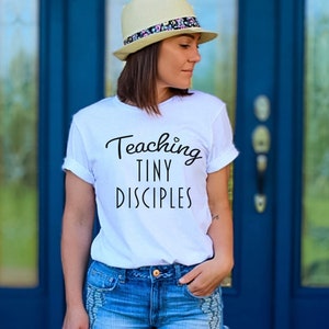 May include: A white t-shirt with the text "Teaching Tiny Disciples" in black lettering. The person wearing the shirt is standing in front of a blue door.
