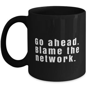 May include: A black ceramic mug with a C-shaped handle featuring the white printed text Go ahead. Blame the network. The mug has a smooth matte finish and a cylindrical body, providing a standard vessel for hot or cold beverages in a simple design.