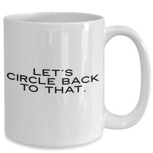 May include: A white ceramic mug featuring the black text LET'S CIRCLE BACK TO THAT printed in the center. The mug has a smooth finish, a cylindrical body, and a curved handle on the right side, providing a standard design for holding beverages.