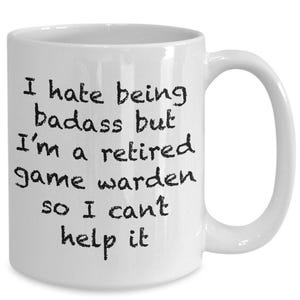 以下が含まれることがあります： 黒い文字で「I hate being badass but I'm a retired game warden so I can't help it」と書かれた白いセラミック製のマグカップ。マグカップは円筒形の本体と右側に湾曲したハンドルを備えており、全体的に滑らかで光沢のある仕上がりになっています。