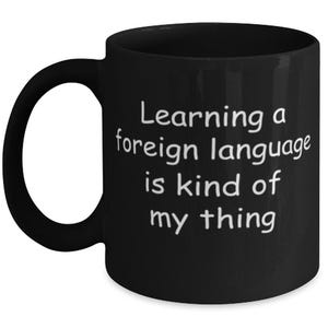 Op de afbeelding: Een zwarte keramische mok met een afgerond handvat en witte bedrukte tekst die luidt: Learning a foreign language is kind of my thing. De mok heeft een gladde afwerking en een standaard cilindrische vorm met een brede opening aan de bovenkant.