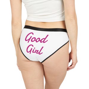 Può includere: Mutandine bianche con rifiniture nere e la scritta "Good Girl" in corsivo rosa. La biancheria intima ha un taglio classico e una fascia in vita nera.
