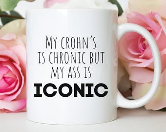 May include: A white ceramic mug featuring black printed text that reads My Crohn's is chronic but my ass is iconic. The mug has a standard cylindrical shape with a curved handle on the right side, presented against a background of soft pink flowers.