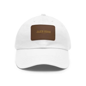 May include: White baseball cap with a curved brim. A brown rectangular patch on the front displays the text "JAKE NINE" in gold lettering. The hat is made of a soft fabric and has a classic, casual style.