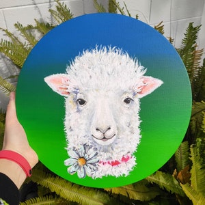 Puede incluir: Un cordero blanco con una flor y una cinta rosa alrededor del cuello está pintado en un lienzo redondo con un fondo degradado de verde a azul.