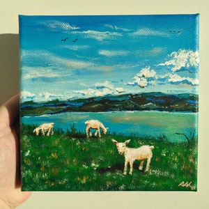 Puede incluir: Una pintura de un paisaje con tres cabras blancas pastando en un campo verde. Las cabras están en primer plano, con un lago azul y montañas en el fondo. El cielo es azul con nubes blancas.