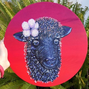 Puede incluir: Una oveja negra con una flor blanca en la cabeza está pintada en un lienzo circular rojo.