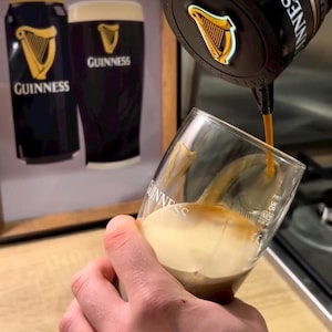 Peut inclure: Verre de Guinness en cours de remplissage. La bi&egrave;re brune coule d'un robinet dans un verre inclin&eacute;. En arri&egrave;re-plan, une image encadr&eacute;e d'une canette et d'un verre de Guinness. Le logo Guinness est visible.