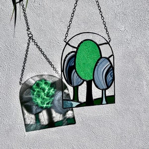 May include: A stained glass window hanging from a chain, depicting three stylized trees with green leaves and blue trunks. The window is casting a shadow on the wall behind it.