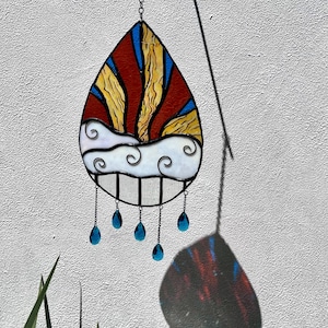 May include: A stained glass suncatcher in the shape of a teardrop with a red, yellow and white design depicting a sunset over clouds. The suncatcher has blue glass teardrop accents hanging from the bottom.